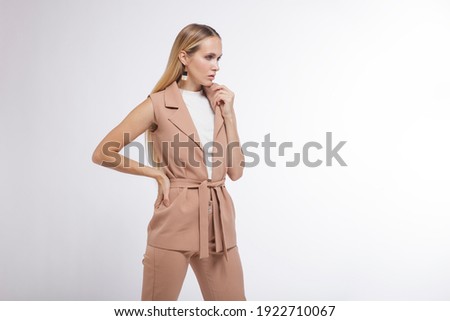 High fashion photo of a beautiful elegant young woman in a pretty beige suit, pants, vest, boots posing over white, soft gray background. Studio Shot, portrait. Blonde