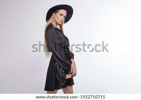 High fashion photo of a beautiful elegant young woman in a pretty black dress, hat, high heels, bag posing over white, soft gray background. Studio Shot, portrait. Blonde