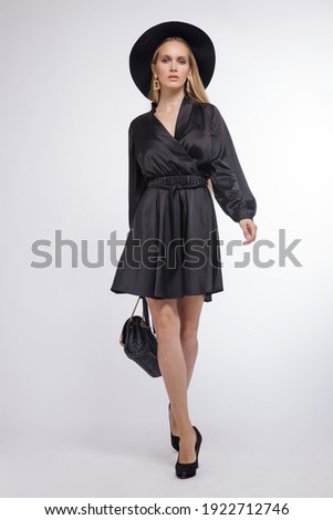 High fashion photo of a beautiful elegant young woman in a pretty black dress, hat, high heels, bag posing over white, soft gray background. Studio Shot, portrait. Blonde