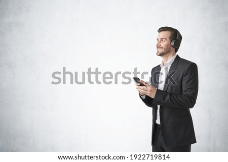 Handsome young European man smiling and listening to music or podcast with his smartphone and headphones over concrete wall background. Concept of relaxation and education, internet. Mock up