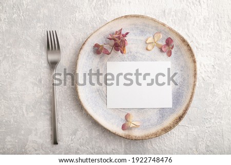 White paper invitation card, mockup with dried hydrangea flowers on ceramic plate and gray concrete background. Blank, flat lay, top view, still life, copy space.