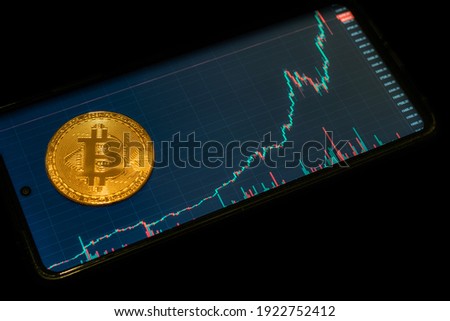 Crypto currency Golden Bitcoin isolated on black background. Photo of coin in front of mobile phone with chart of bitcoin value. Blockchain technology, bitcoin mining concept.