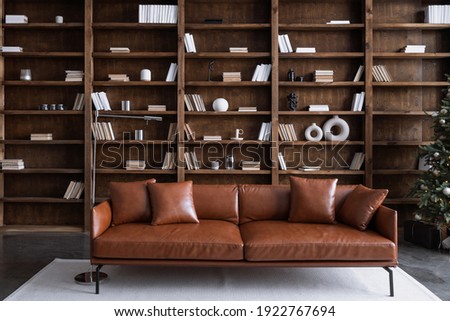 Leather sofa with cushions standing on living room with stylish interior design and collections books on bookshelves in library. Work cabinet in modern apartment