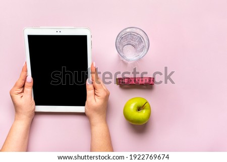 girl holds tablet with black screen in her hands, green apple, measuring tape, bottle of water on pink background Top view Flat lay Mock up Healthy lifestyle, sports, diet blog concept