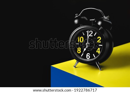 The hands of the alarm clock indicate seven o'clock. A black round alarm clock stands on the corner of a yellow-blue cube. Background with space to copy.