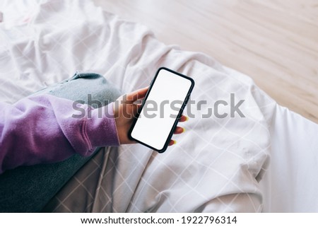 Woman holding a smartphone with a white screen mock up, resting on the bed at home.