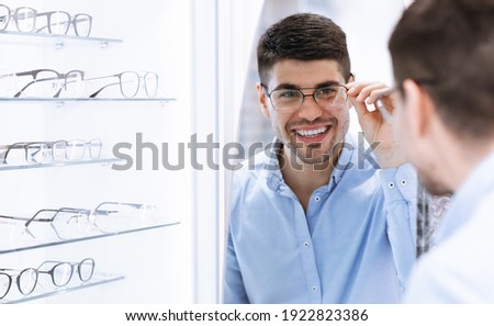 Satisfied Customer. Over the shoulder view of happy young male client wearing new glasses, looking in the mirror standing near rack and showcase with eyewear. Smiling man trying on spectacles Royalty-Free Stock Photo #1922823386