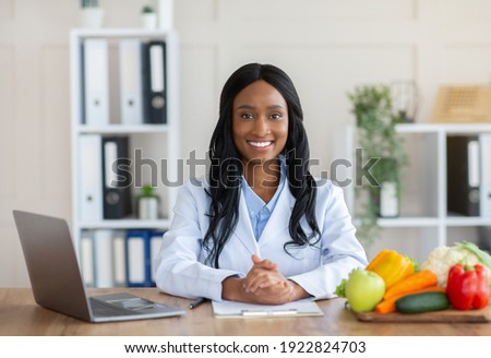 Smart eating and healthy nutrition concept. Portrait of positive black dietitian looking at camera and smiling at workplace. Young African American nutritionist giving online weight loss consultations Royalty-Free Stock Photo #1922824703