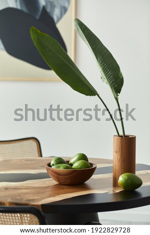 Minimalist composition on the design wooden table with fruits, tropical leaf in vase, abstract painitngs and stylish chair. Modern dining room. Template. 