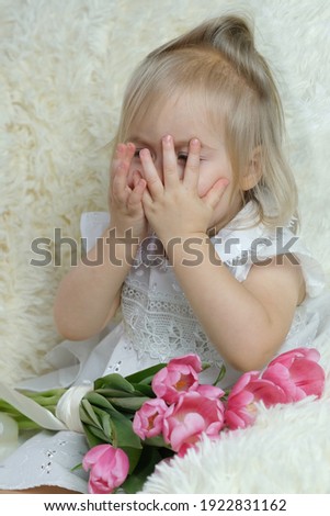 girl of two years old, blonde is sitting in the room on the couch with a bouquet of pink tulips, covered her eyes with her hands, concept small woman, birthday, happy childhood