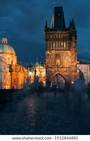 Blurred ghost like people silhouettes walking on illuminated Charles Bridge in Prague in night on Gothic towers background