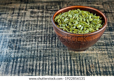 Peeled pumpkin seeds in a clay bowl on a wooden table. Seeds on a black shabby board. Copy space and free space for text near the seeds.