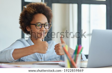 Enthusiastic mixed race teen schoolgirl in glasses showing thumbs up to her teacher during online video lesson while studying from home