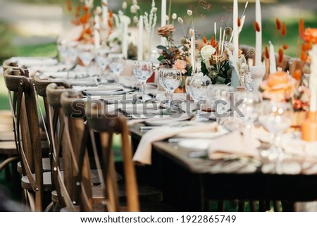 Boho wedding table for guests at bouquet after wedding ceremony and photo for marriage blog about interior. Candles, bouquets, napkins, dishes, accessories and glasses on table with chairs, outdoors
