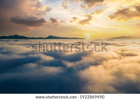 Aerial view of vibrant sunset over white dense clouds with distant dark mountains on horizon. Royalty-Free Stock Photo #1922869490