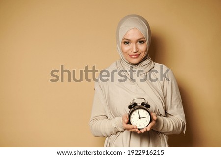 Cheerful young muslim woman in hijab holding alarm clock in hands and smiling cute at camera standing against a beige background with copy space.