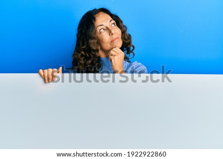 Middle age hispanic woman holding blank empty banner with hand on chin thinking about question, pensive expression. smiling with thoughtful face. doubt concept. 