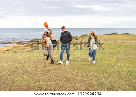 Three mixed-race carefree friends running outdoor joking each other - Millennial people having fun on a meadow near sea smiling in winter vacation enjoying nature in ocean resort wearing clothing