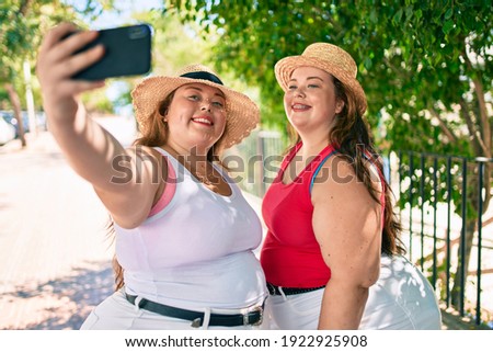 Two plus size overweight sisters twins women smiling taking a selfie picture with the phone outdoors on a sunny day