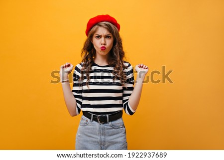 Positive woman in striped shirt and beret makes funny face. Curly young lady in denim skirt with wide black belt posing on isolated background..