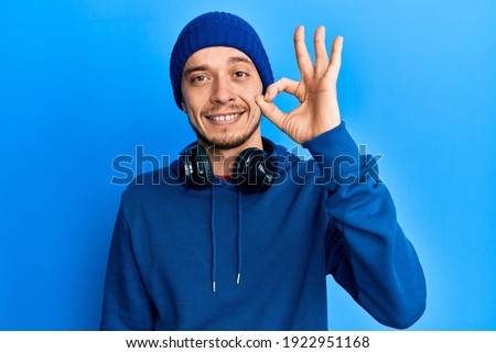 Hispanic young man wearing sweatshirt and headphones smiling positive doing ok sign with hand and fingers. successful expression. 
