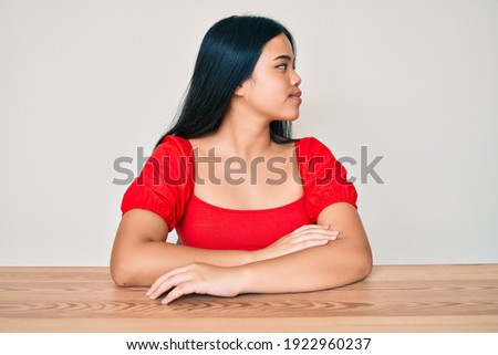 Young beautiful asian girl wearing casual clothes sitting on the table looking to side, relax profile pose with natural face with confident smile. 