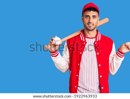 Young hispanic man playing baseball holding bat and ball smiling happy pointing with hand and finger to the side 