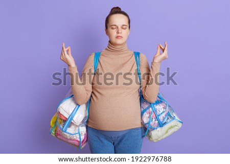Calming pregnant woman isolated over lilac background, keeps eyes closed, meditates, trying to relax, wearing casual clothing, expectant mother holding bags with stuff.