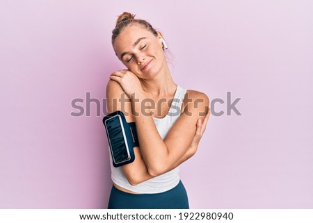 Beautiful blonde sport woman wearing arm band and earphones hugging oneself happy and positive, smiling confident. self love and self care 