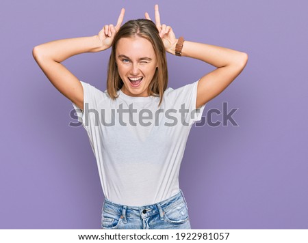 Beautiful young blonde woman wearing casual white t shirt posing funny and crazy with fingers on head as bunny ears, smiling cheerful 