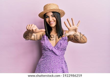 Young brunette woman wearing summer dress and hat showing and pointing up with fingers number six while smiling confident and happy. 