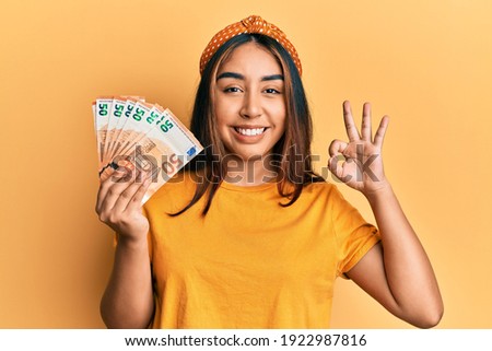 Young latin woman holding 50 euro banknotes doing ok sign with fingers, smiling friendly gesturing excellent symbol 