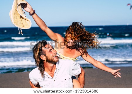 beautiful couple of adults having fun together at the beach on the sand - man walking with his wife or his girlfriend on he - he's holding she - woman with arms open be happy