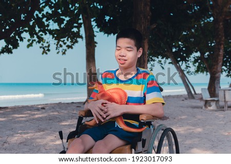 Asian disabled child on wheelchair hug a dinosaur on summer of sea beach nature background,Lifestyle of special child in the education age,Happy disabled kid concept,Toy is best friend in alone mood.