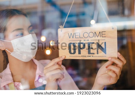 Smiling young asian owner retail,coffee shop woman turning sign board to open wearing face mask,protect to pandemic of coronavirus, reopen store after close lockdown quarantine in covid to new normal.