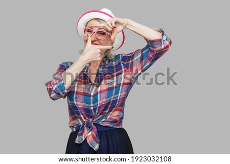 Portrait of attentive modern stylish mature woman in casual style with hat and eyeglasses standing with crop composition gesture and looking at target. indoor studio shot isolated on gray background