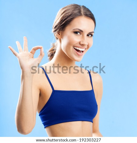 Cheerful woman in fitness wear showing okay gesture, over blue background