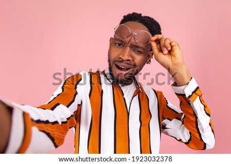 What did you say? Surprised confused afro-american man blogger with dreadlocks looking at selfie camera, chatting with subscribers online, streaming. Indoor studio shot isolated on pink background