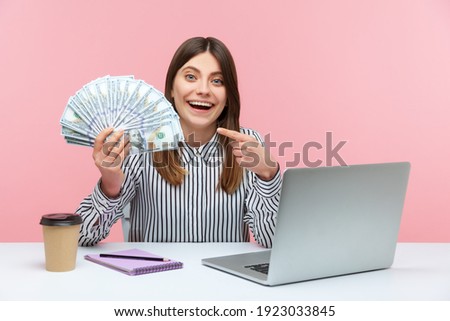 Excited positive business woman pointing finger at hundred dollar bills she holding sitting at workplace with laptop, high salary, bonuses and perks. Indoor studio shot isolated on pink background Royalty-Free Stock Photo #1923033845