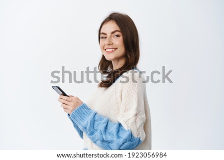 Portrait of beautiful female model using smartphone app, standing in profile and turn face with smile at camera, sending message in social media chat, white background.