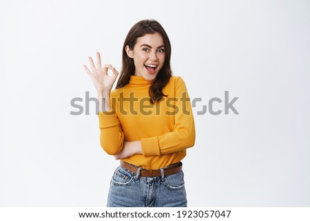 Very good. Smiling young female model with natural make up, showing okay sign and say yes, confirm product is alright, approve and like something, standing on white background.