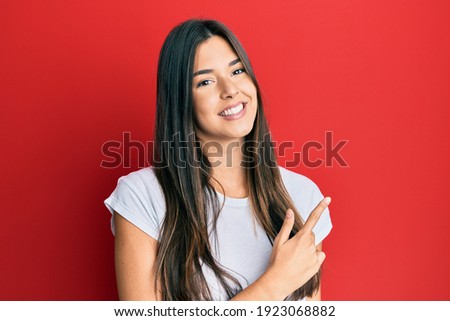 Young brunette woman wearing casual white tshirt over red background cheerful with a smile of face pointing with hand and finger up to the side with happy and natural expression on face 