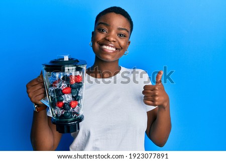 Young african american woman holding food processor mixer machine with fruits smiling happy and positive, thumb up doing excellent and approval sign 