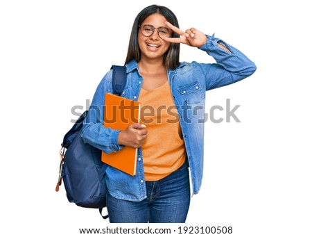 Young latin girl wearing student backpack and holding books doing peace symbol with fingers over face, smiling cheerful showing victory 