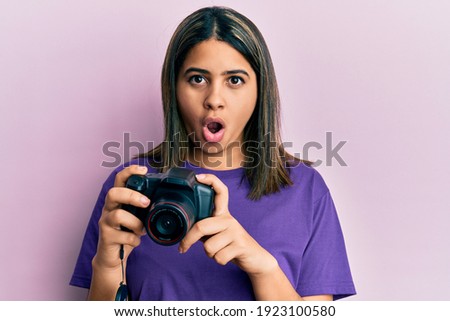 Young latin woman using reflex camera afraid and shocked with surprise and amazed expression, fear and excited face. 