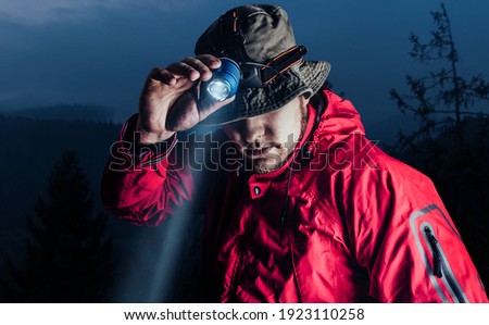 Photo of a male hiker in red jacket and panama putting on head mount flashlight on dark mountain forest background. Royalty-Free Stock Photo #1923110258