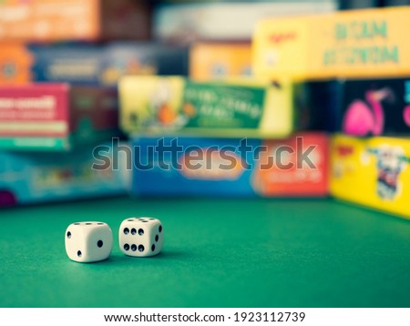 White dice on the green surface on the blurred background of colorful board game boxes. Concept of entertainment under lockdown