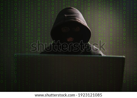 A dangerous masked hooded hacker infiltrates data servers and infects their systems with viruses. Male criminal steals information through a laptop. Photo on a dark background in the studio