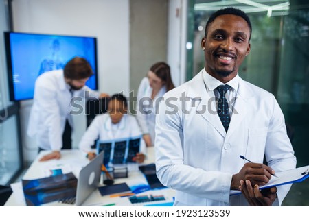 African American doctor looking at the camera. behind him, doctors of different nationalities are talking, sitting at the table in the first-aid post. Royalty-Free Stock Photo #1923123539