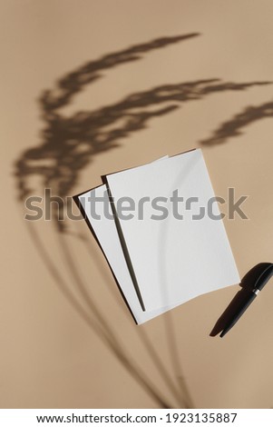 A blank greeting card, desaturated mock up. An empty sheet of white paper on a pastel yellow surface, a black pen and a shadow of wheat spikes  